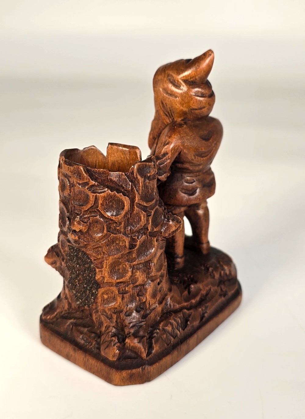 A carved brown limewood match holder depicting a hooded bearded gnome standing beside a textured tree stump container. View IV