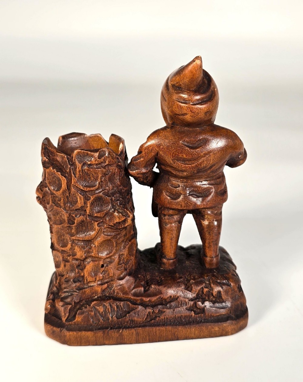 A carved brown limewood match holder depicting a hooded bearded gnome standing beside a textured tree stump container. View V