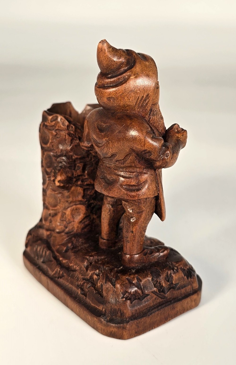 A carved brown limewood match holder depicting a hooded bearded gnome standing beside a textured tree stump container. View VI