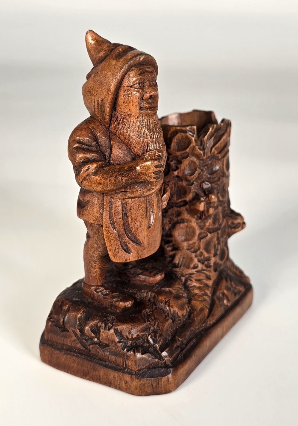 A carved brown limewood match holder depicting a hooded bearded gnome standing beside a textured tree stump container. View VII