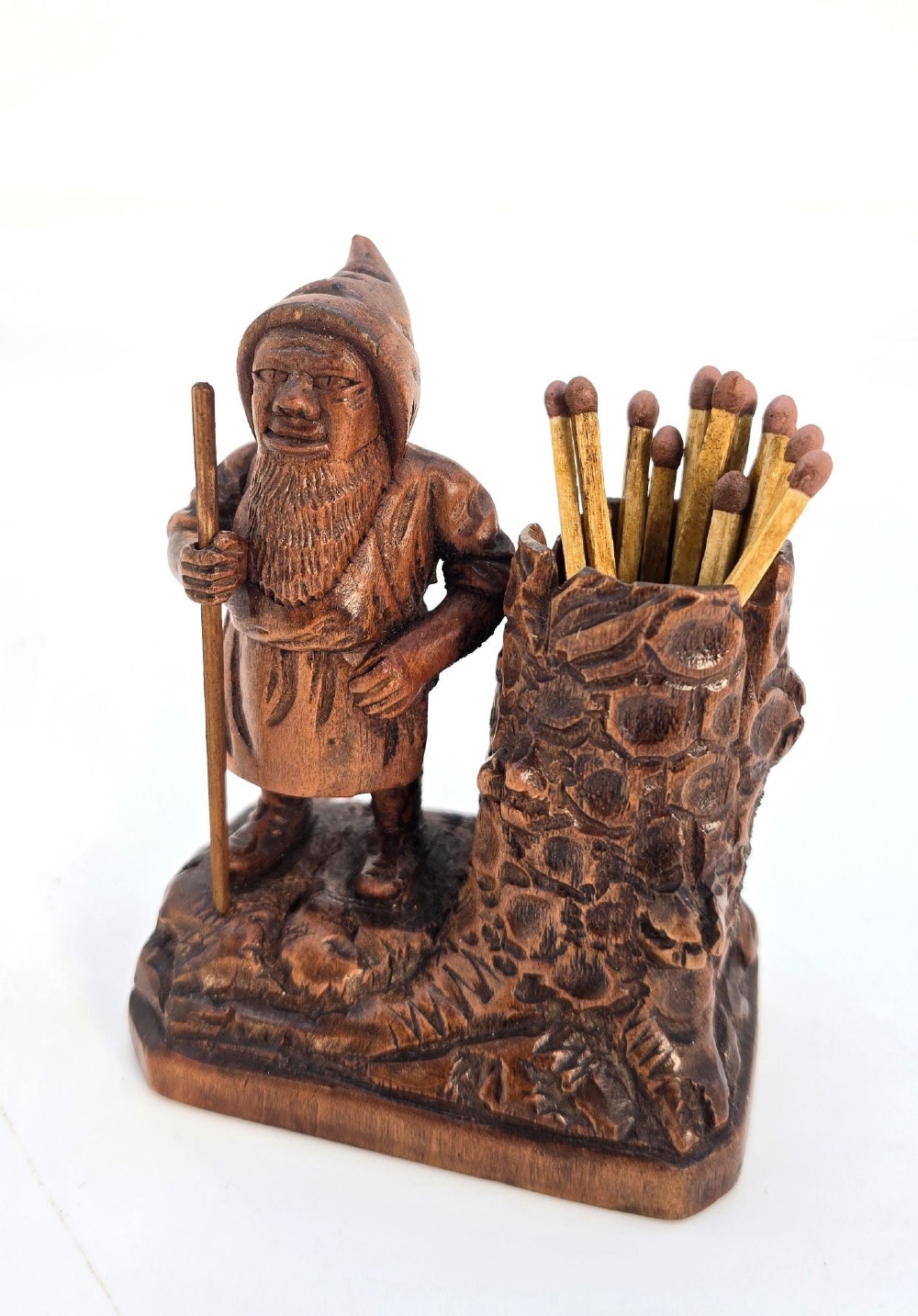 A carved brown limewood match holder depicting a hooded bearded gnome standing beside a textured tree stump container. View VIII