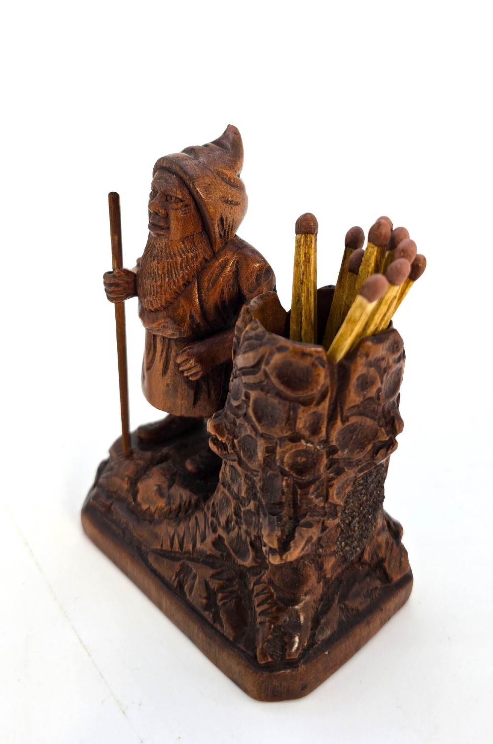 A carved brown limewood match holder depicting a hooded bearded gnome standing beside a textured tree stump container. View IX