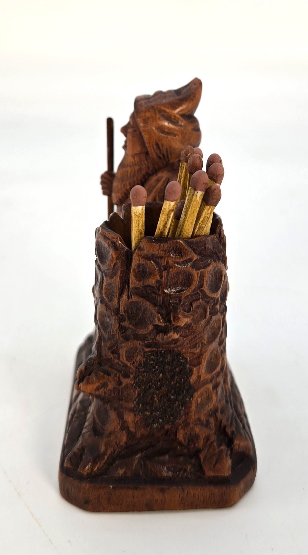 A carved brown limewood match holder depicting a hooded bearded gnome standing beside a textured tree stump container. View X