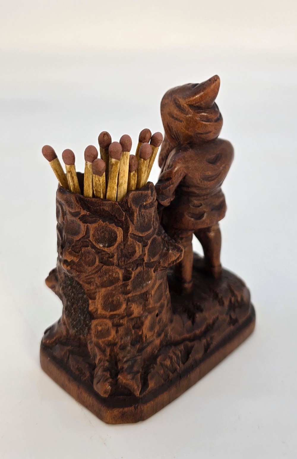 A carved brown limewood match holder depicting a hooded bearded gnome standing beside a textured tree stump container. View XI