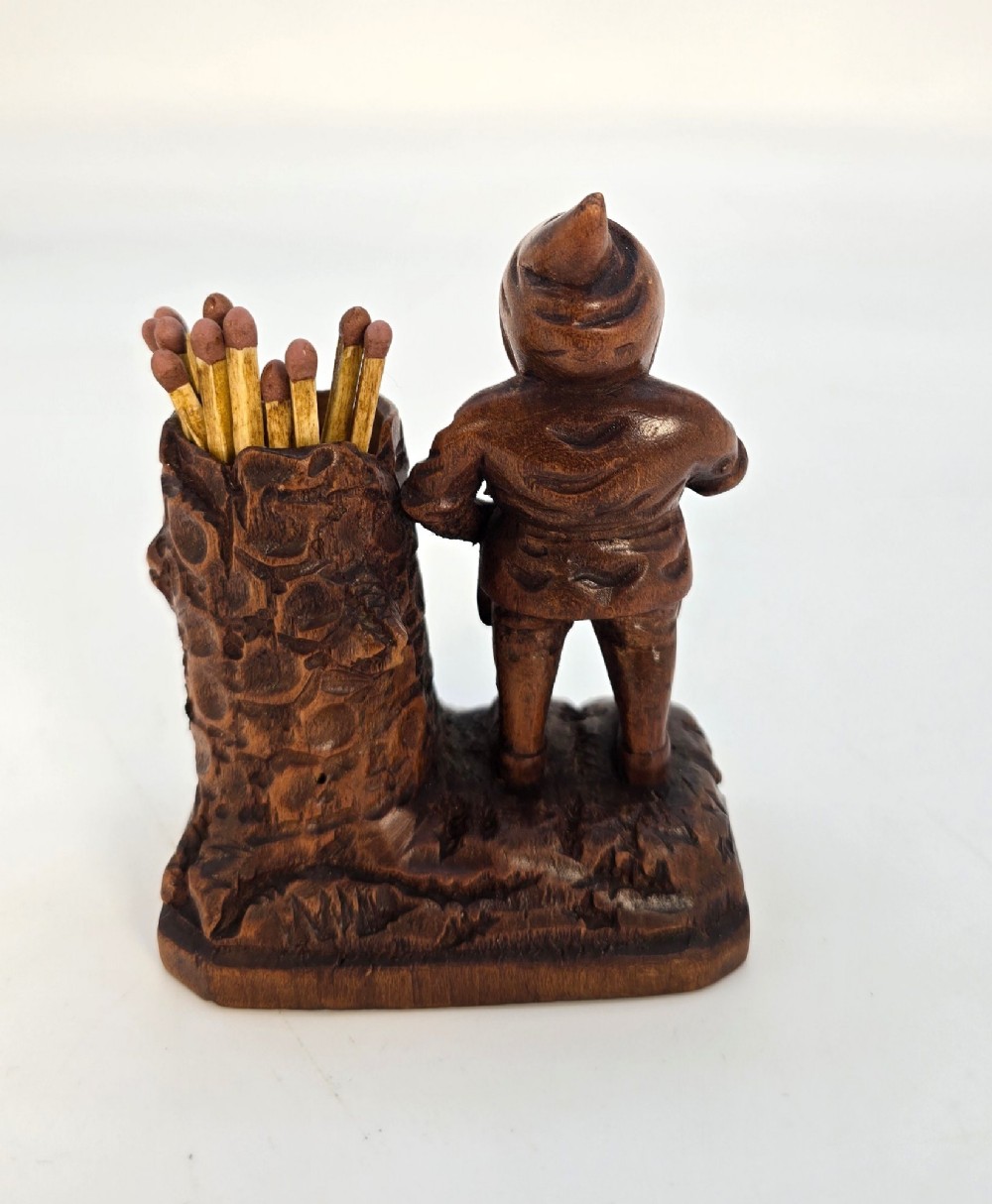 A carved brown limewood match holder depicting a hooded bearded gnome standing beside a textured tree stump container. View XII