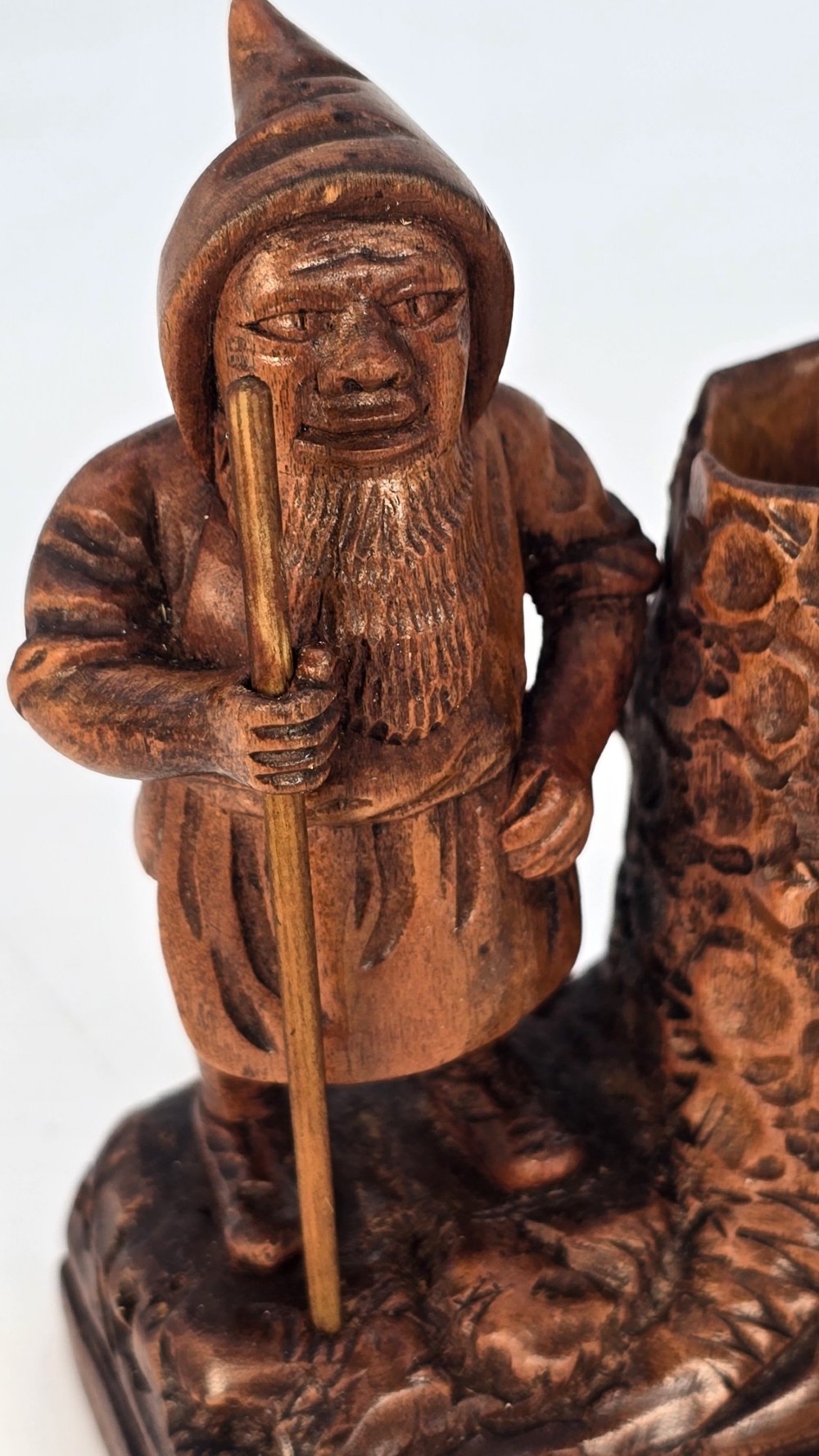 A carved brown limewood match holder depicting a hooded bearded gnome standing beside a textured tree stump container. View XV