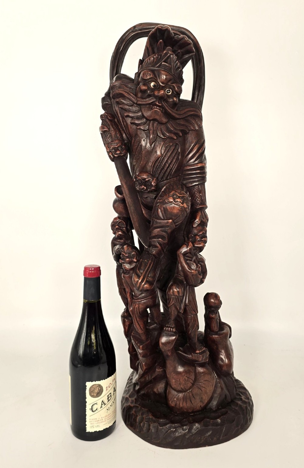 A tall carved dark hardwood Chinese sculpture depicting a Guardian King wearing detailed armour and holding a sword over attendant figures. View II