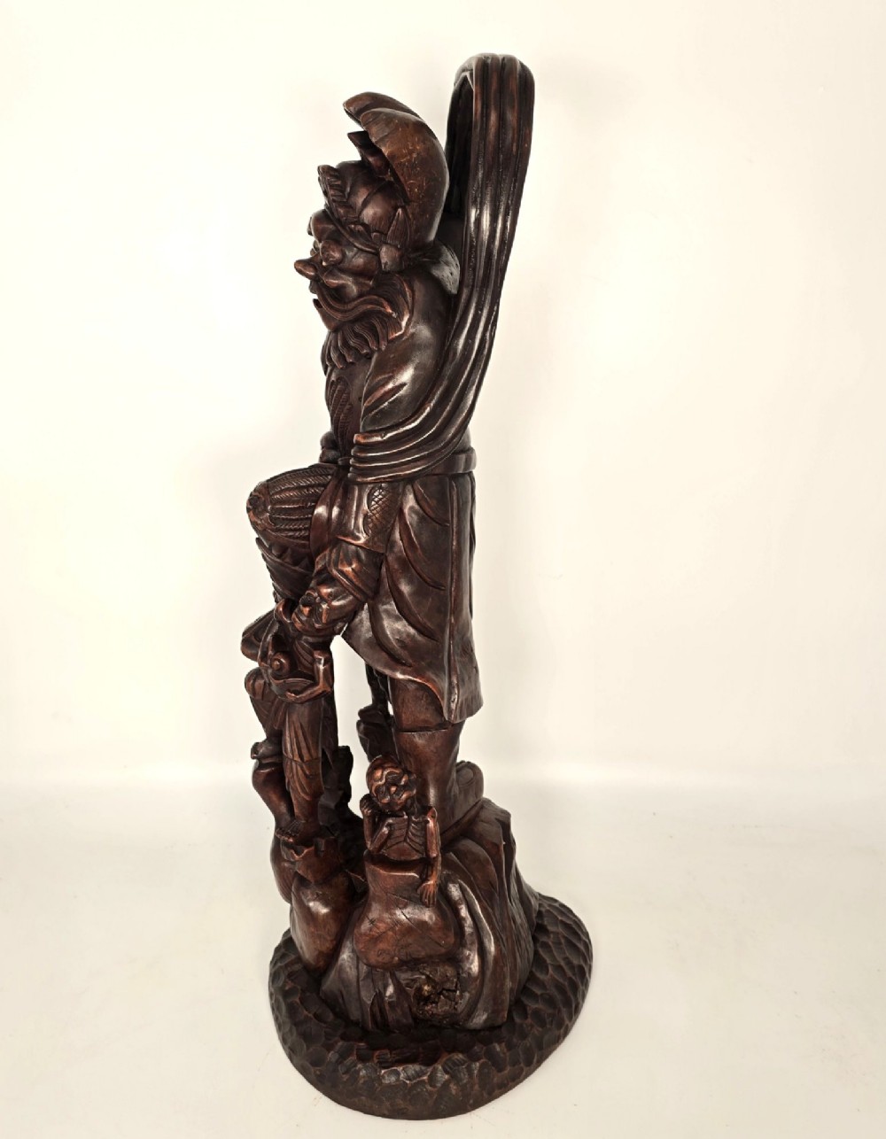 A tall carved dark hardwood Chinese sculpture depicting a Guardian King wearing detailed armour and holding a sword over attendant figures. View VII