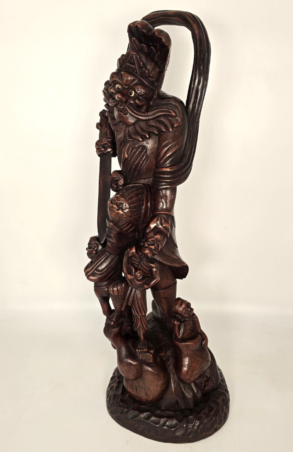 A tall carved dark hardwood Chinese sculpture depicting a Guardian King wearing detailed armour and holding a sword over attendant figures. View VIII