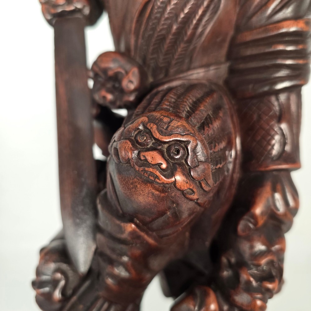A tall carved dark hardwood Chinese sculpture depicting a Guardian King wearing detailed armour and holding a sword over attendant figures. View X