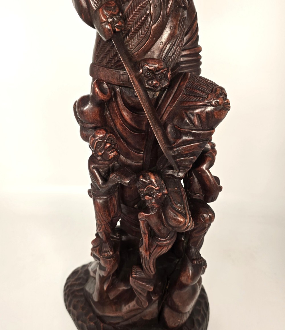 A tall carved dark hardwood Chinese sculpture depicting a Guardian King wearing detailed armour and holding a sword over attendant figures. View XII