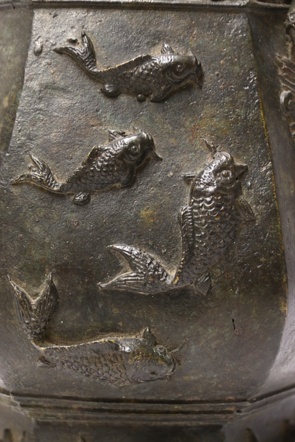 An antique hexagonal solid bronze vessel featuring relief carvings of fish, architectural structures, floral bands, and twin animal-head ring handles. View IV
