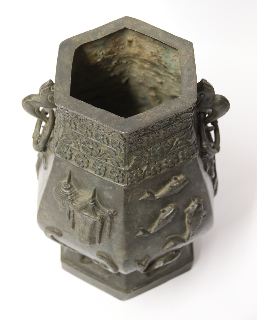 An antique hexagonal solid bronze vessel featuring relief carvings of fish, architectural structures, floral bands, and twin animal-head ring handles. View XIV