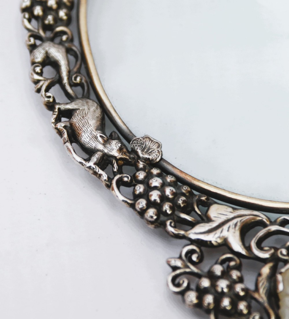 An antique Chinese magnifying glass featuring an ornate repousse silver circular frame decorated with squirrels and grapevines attached to a carved off-white jade handle. View II