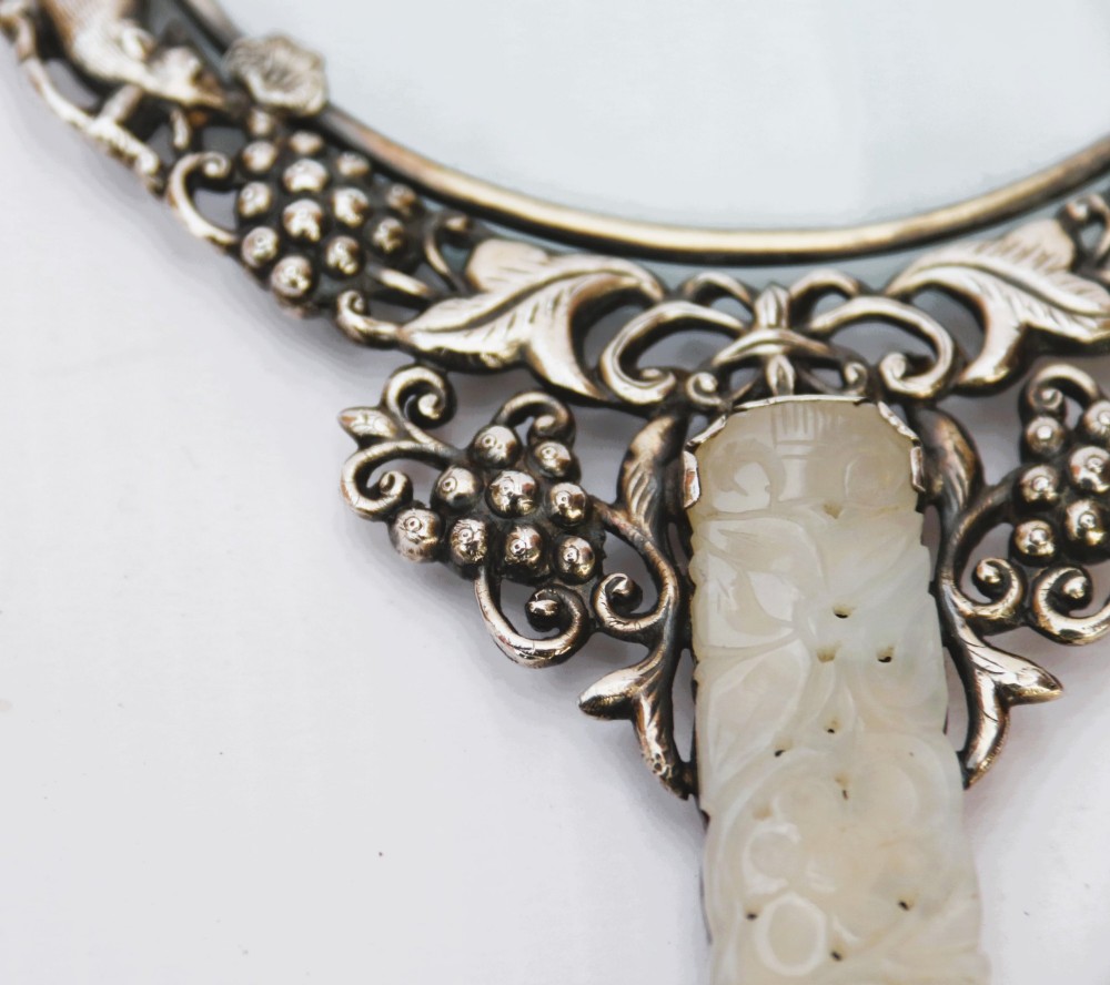 An antique Chinese magnifying glass featuring an ornate repousse silver circular frame decorated with squirrels and grapevines attached to a carved off-white jade handle. View V