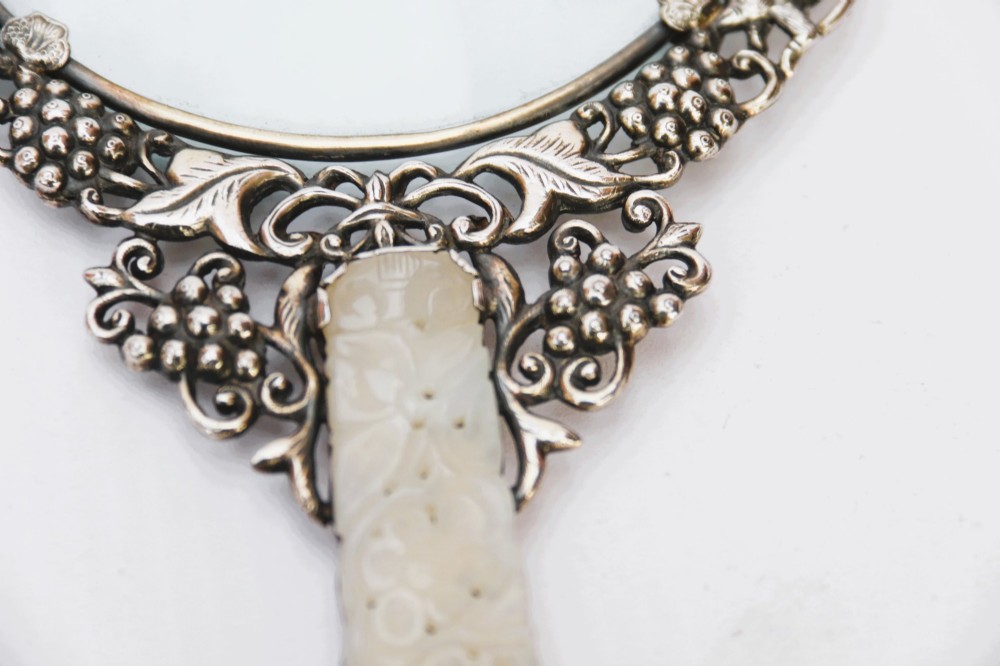 An antique Chinese magnifying glass featuring an ornate repousse silver circular frame decorated with squirrels and grapevines attached to a carved off-white jade handle. View VI