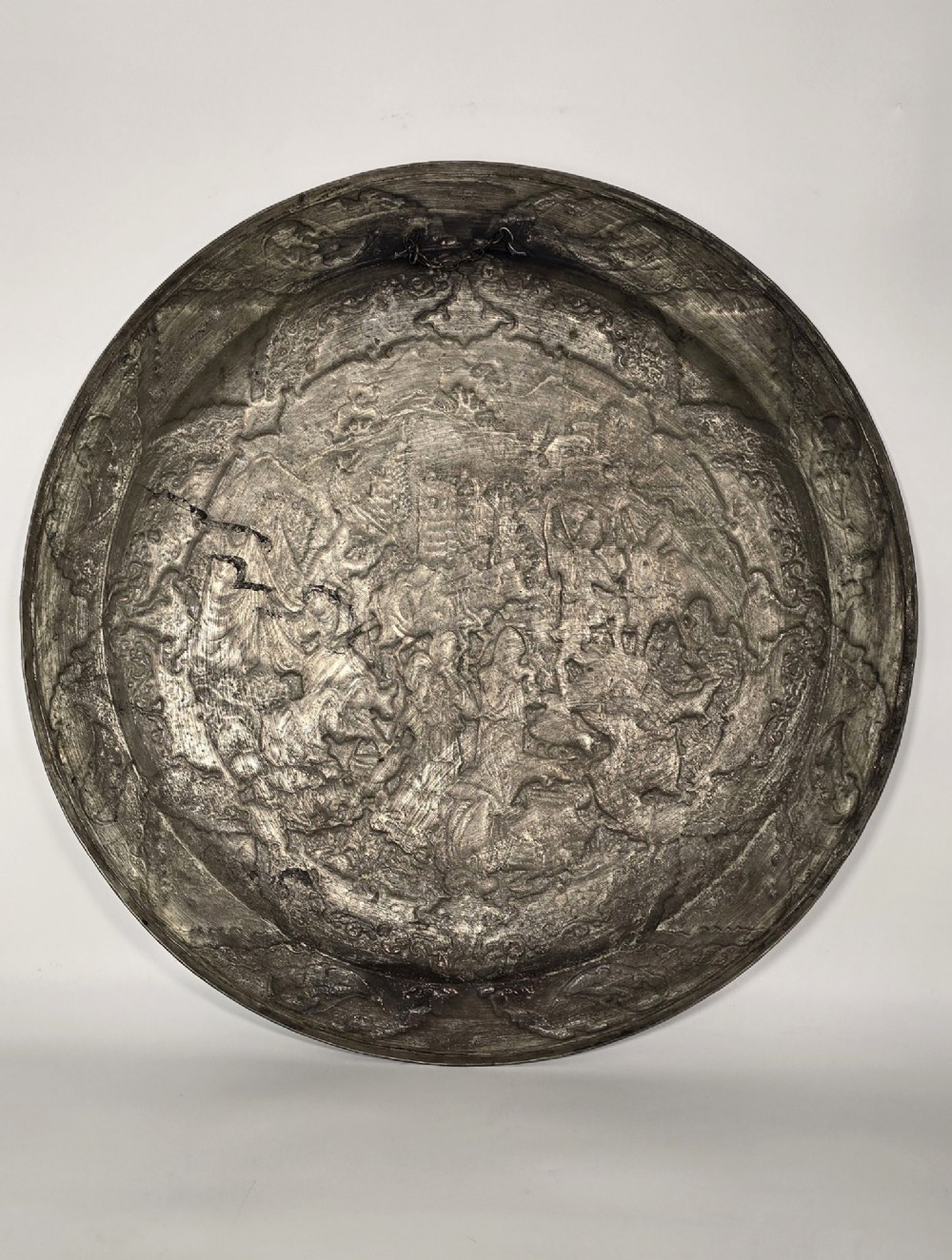 A circular 19th-century Persian copper charger plate featuring dense repousse and engraved figural and landscape scenes surrounded by ornate floral borders. View XII