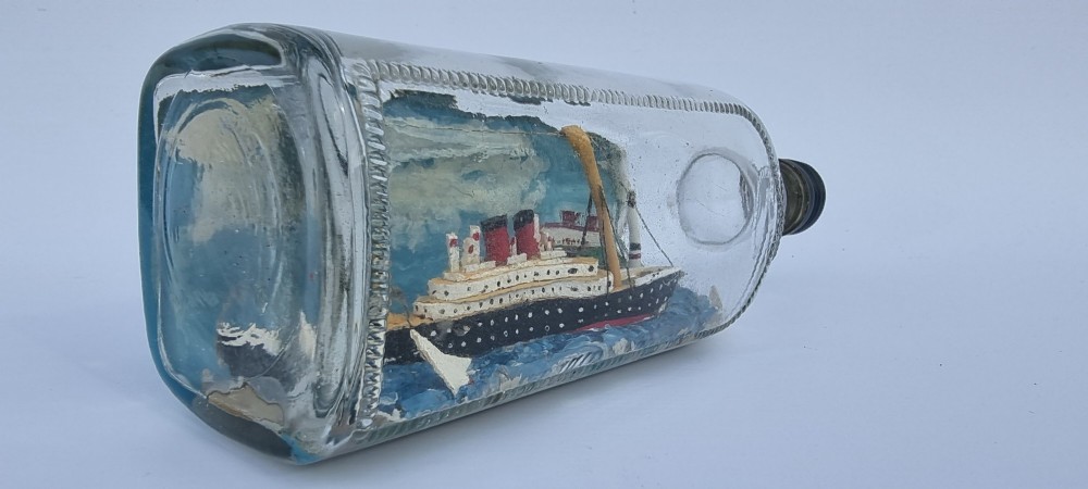 A horizontal glass bottle containing a handcrafted 1930s steamship diorama with red funnels, a painted blue sea, and a lighthouse background. View II