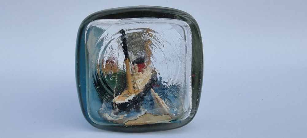 A horizontal glass bottle containing a handcrafted 1930s steamship diorama with red funnels, a painted blue sea, and a lighthouse background. View III