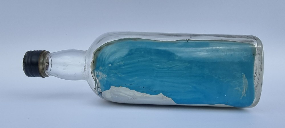 A horizontal glass bottle containing a handcrafted 1930s steamship diorama with red funnels, a painted blue sea, and a lighthouse background. View IV