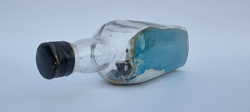 A horizontal glass bottle containing a handcrafted 1930s steamship diorama with red funnels, a painted blue sea, and a lighthouse background. View V
