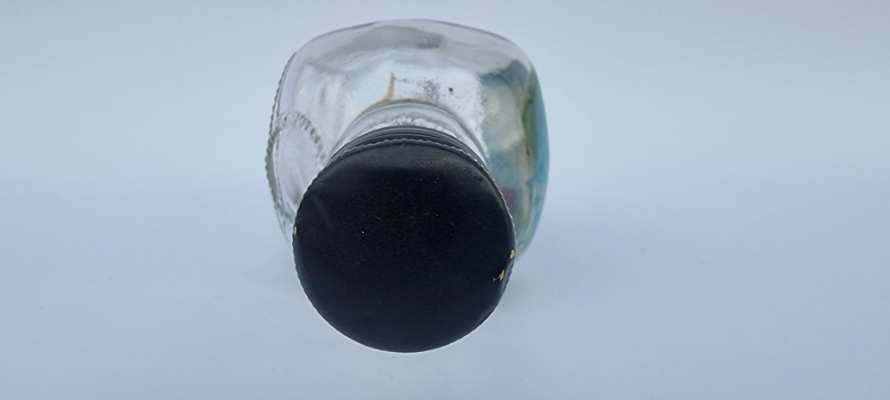 A horizontal glass bottle containing a handcrafted 1930s steamship diorama with red funnels, a painted blue sea, and a lighthouse background. View VI