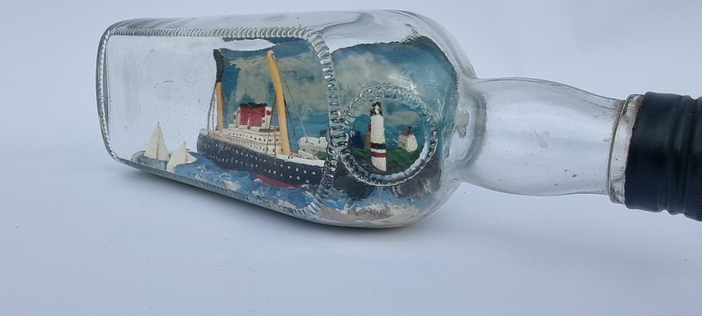 A horizontal glass bottle containing a handcrafted 1930s steamship diorama with red funnels, a painted blue sea, and a lighthouse background. View VII