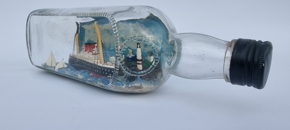 A horizontal glass bottle containing a handcrafted 1930s steamship diorama with red funnels, a painted blue sea, and a lighthouse background. View VIII