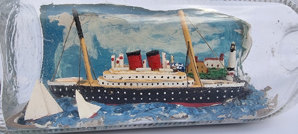 A horizontal glass bottle containing a handcrafted 1930s steamship diorama with red funnels, a painted blue sea, and a lighthouse background. View IX