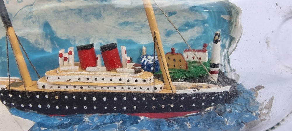 A horizontal glass bottle containing a handcrafted 1930s steamship diorama with red funnels, a painted blue sea, and a lighthouse background. View X
