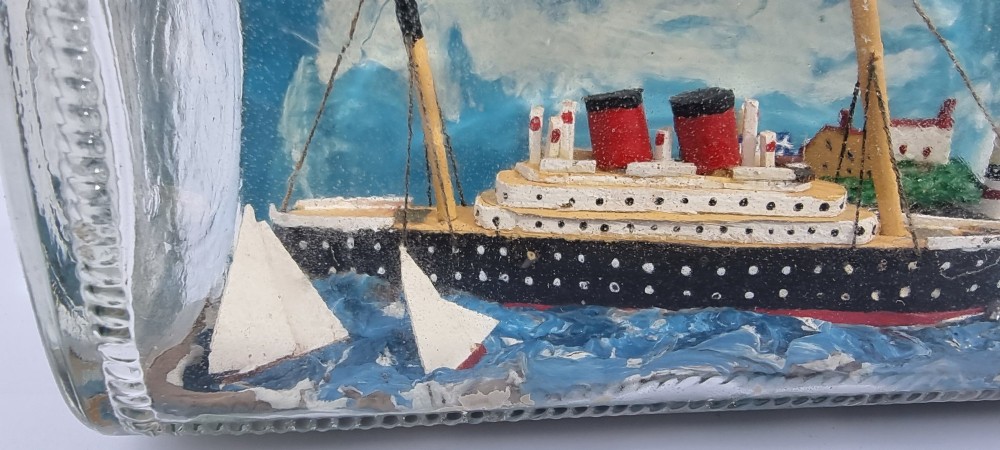 A horizontal glass bottle containing a handcrafted 1930s steamship diorama with red funnels, a painted blue sea, and a lighthouse background. View XI