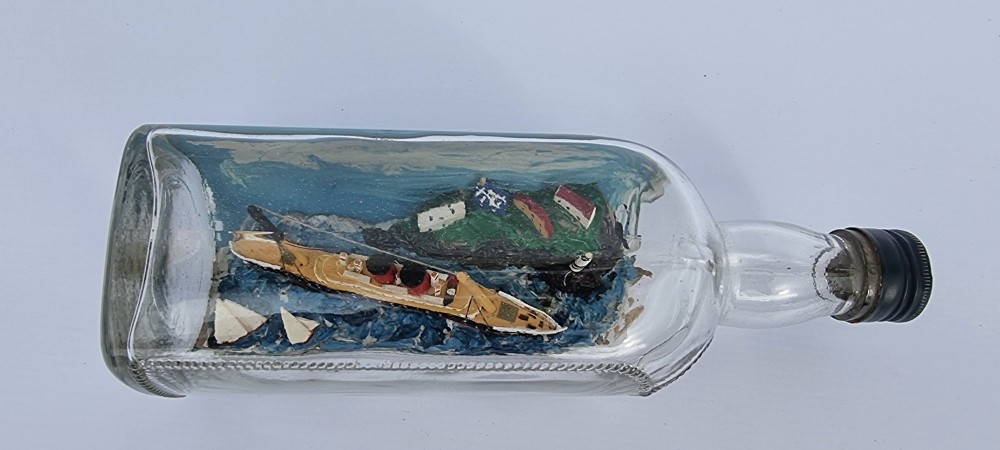 A horizontal glass bottle containing a handcrafted 1930s steamship diorama with red funnels, a painted blue sea, and a lighthouse background. View XII