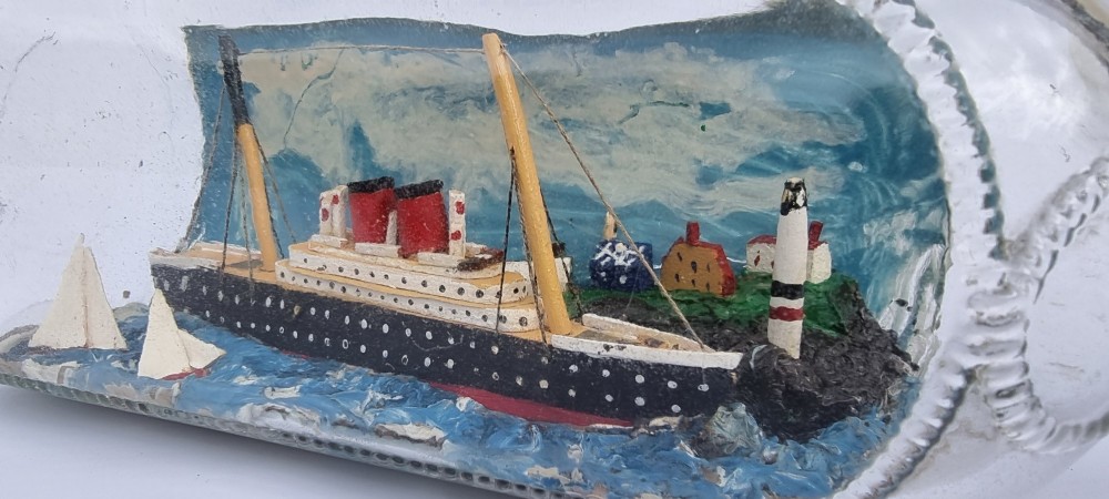 A horizontal glass bottle containing a handcrafted 1930s steamship diorama with red funnels, a painted blue sea, and a lighthouse background. View XIII
