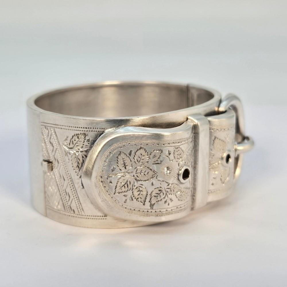 A wide Victorian sterling silver hinged bangle crafted with a functional buckle motif and detailed hand-engraved foliage patterns.