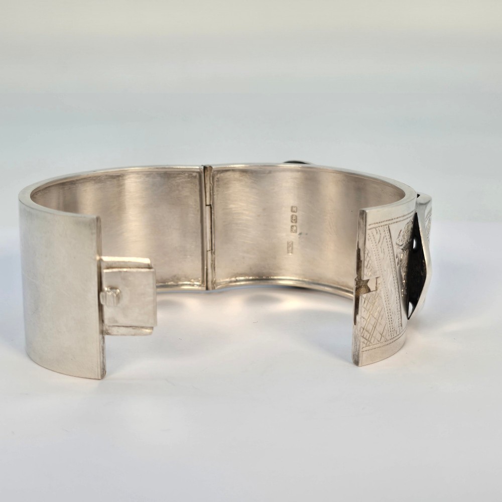 A wide Victorian sterling silver hinged bangle crafted with a functional buckle motif and detailed hand-engraved foliage patterns. View VI