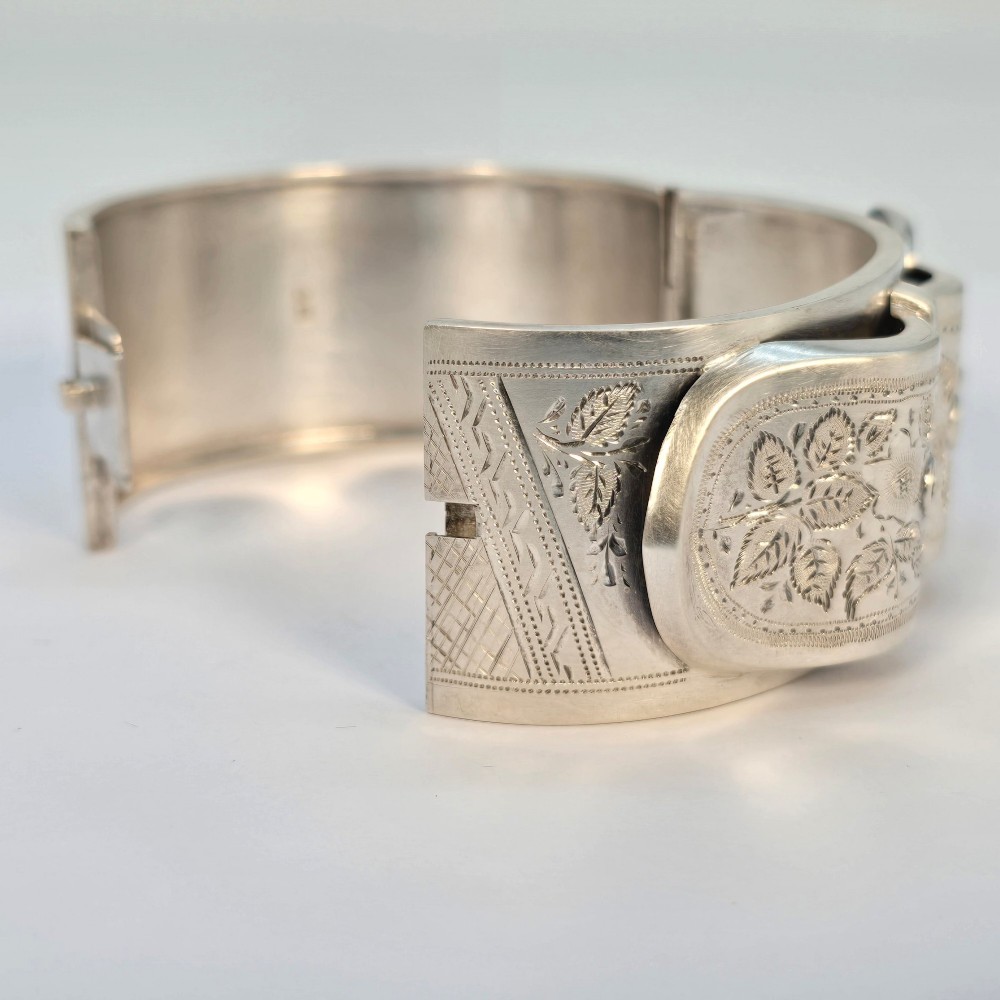 A wide Victorian sterling silver hinged bangle crafted with a functional buckle motif and detailed hand-engraved foliage patterns. View VII