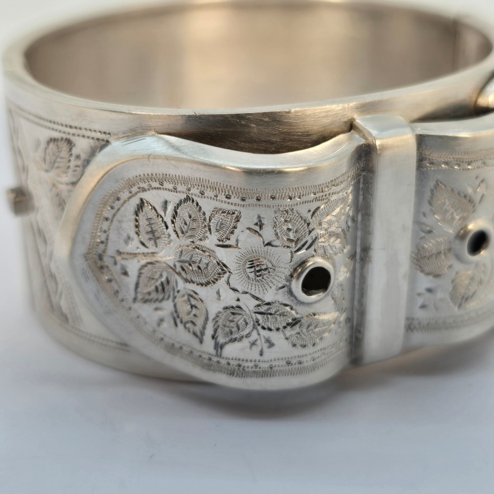 A wide Victorian sterling silver hinged bangle crafted with a functional buckle motif and detailed hand-engraved foliage patterns. View IX