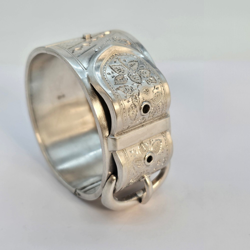 A wide Victorian sterling silver hinged bangle crafted with a functional buckle motif and detailed hand-engraved foliage patterns. View X