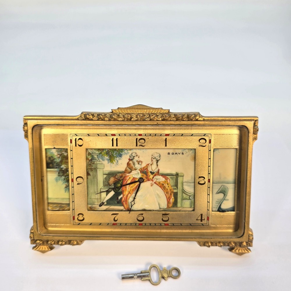 A rectangular gilt brass Art Deco strut clock featuring an illustrated courtly scene on the central dial surrounded by numeric markers.