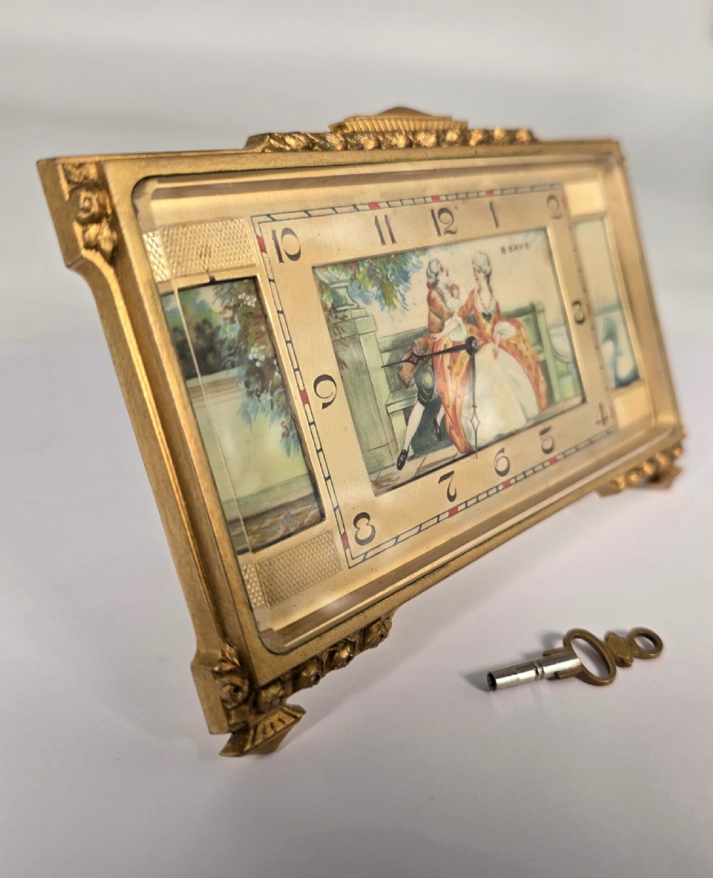 A rectangular gilt brass Art Deco strut clock featuring an illustrated courtly scene on the central dial surrounded by numeric markers. View II