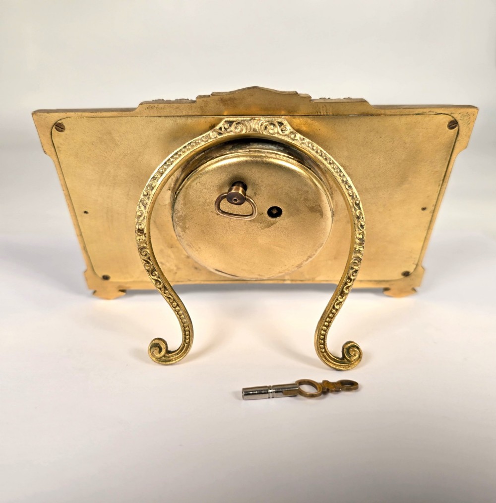 A rectangular gilt brass Art Deco strut clock featuring an illustrated courtly scene on the central dial surrounded by numeric markers. View IV