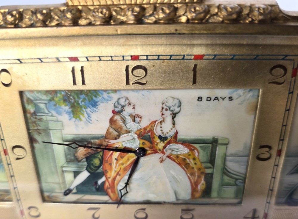 A rectangular gilt brass Art Deco strut clock featuring an illustrated courtly scene on the central dial surrounded by numeric markers. View VI