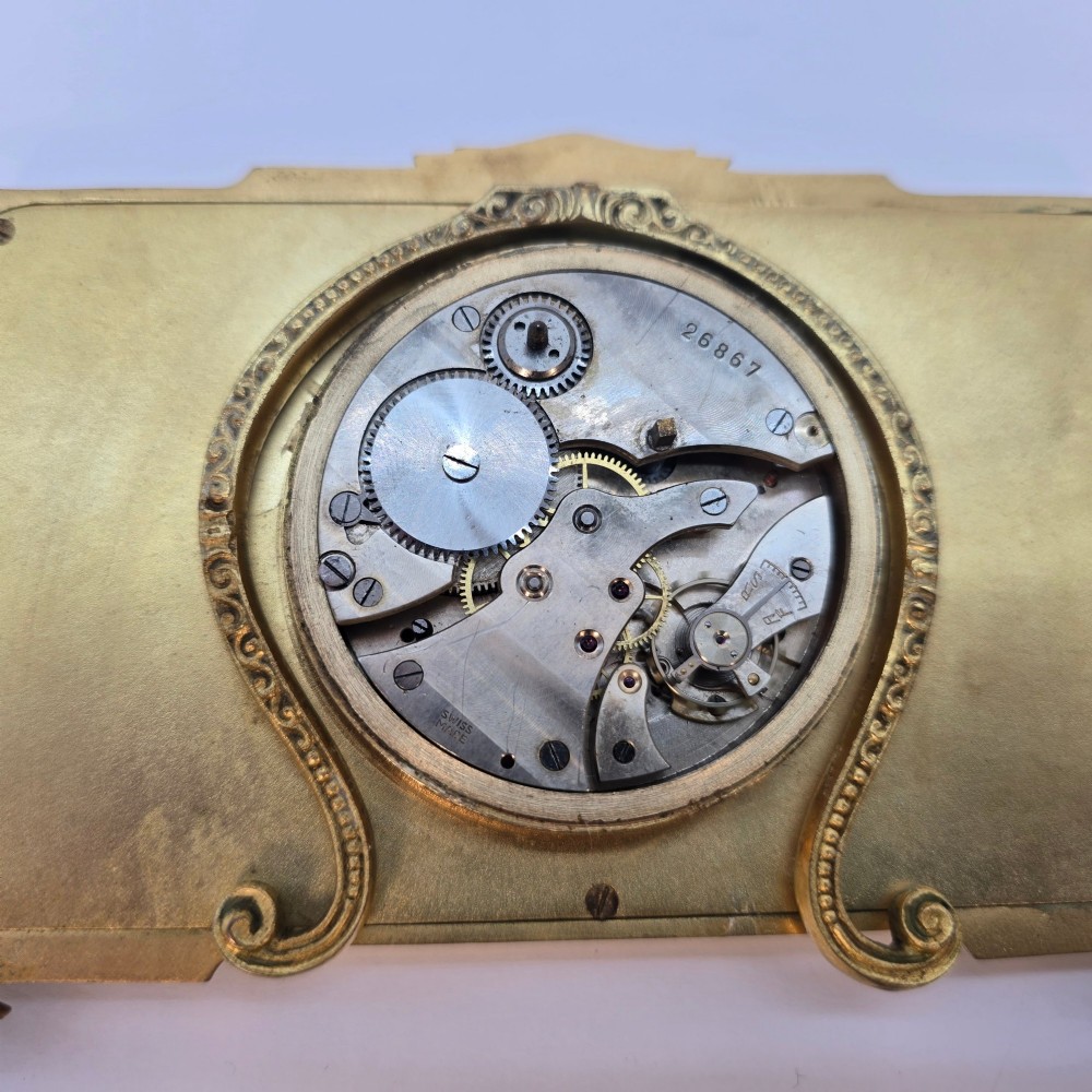 A rectangular gilt brass Art Deco strut clock featuring an illustrated courtly scene on the central dial surrounded by numeric markers. View XII