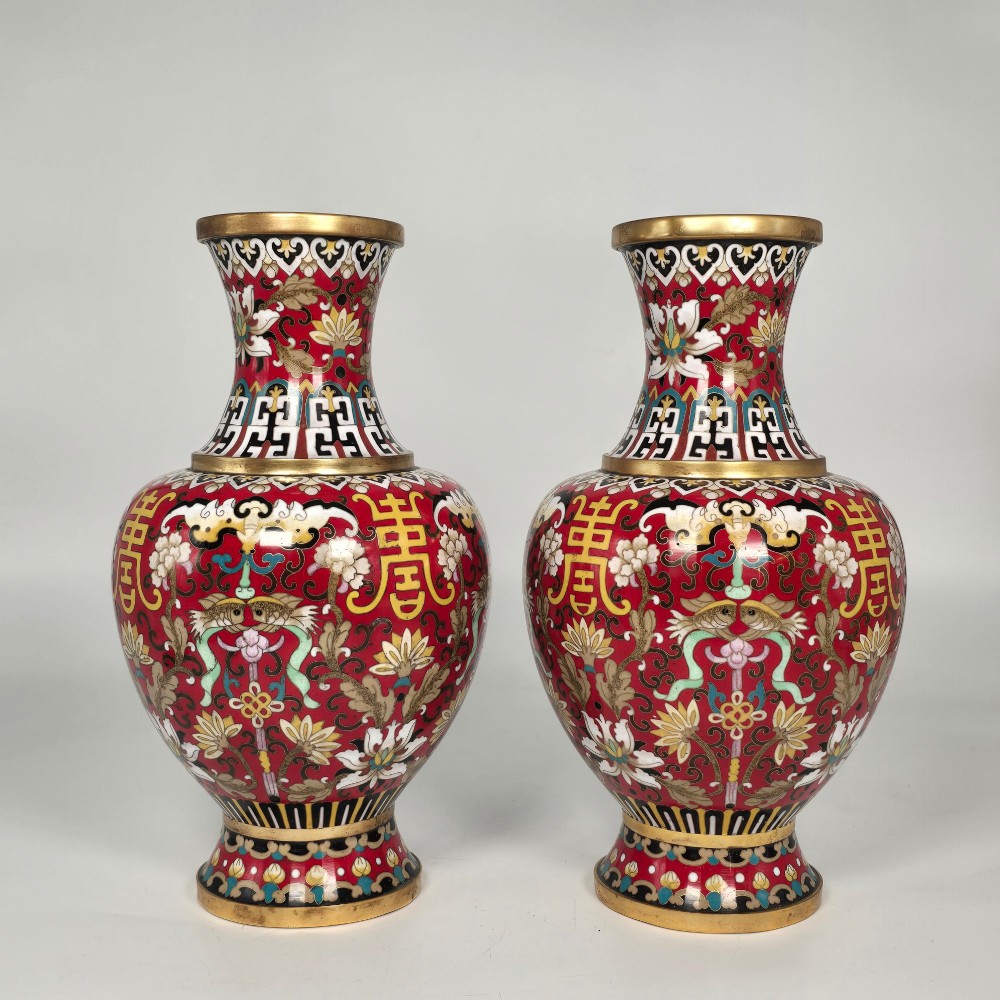 A matching pair of baluster-form Chinese cloisonne enamel vases with red backgrounds, lotus motifs, auspicious symbols, and gilt rims.