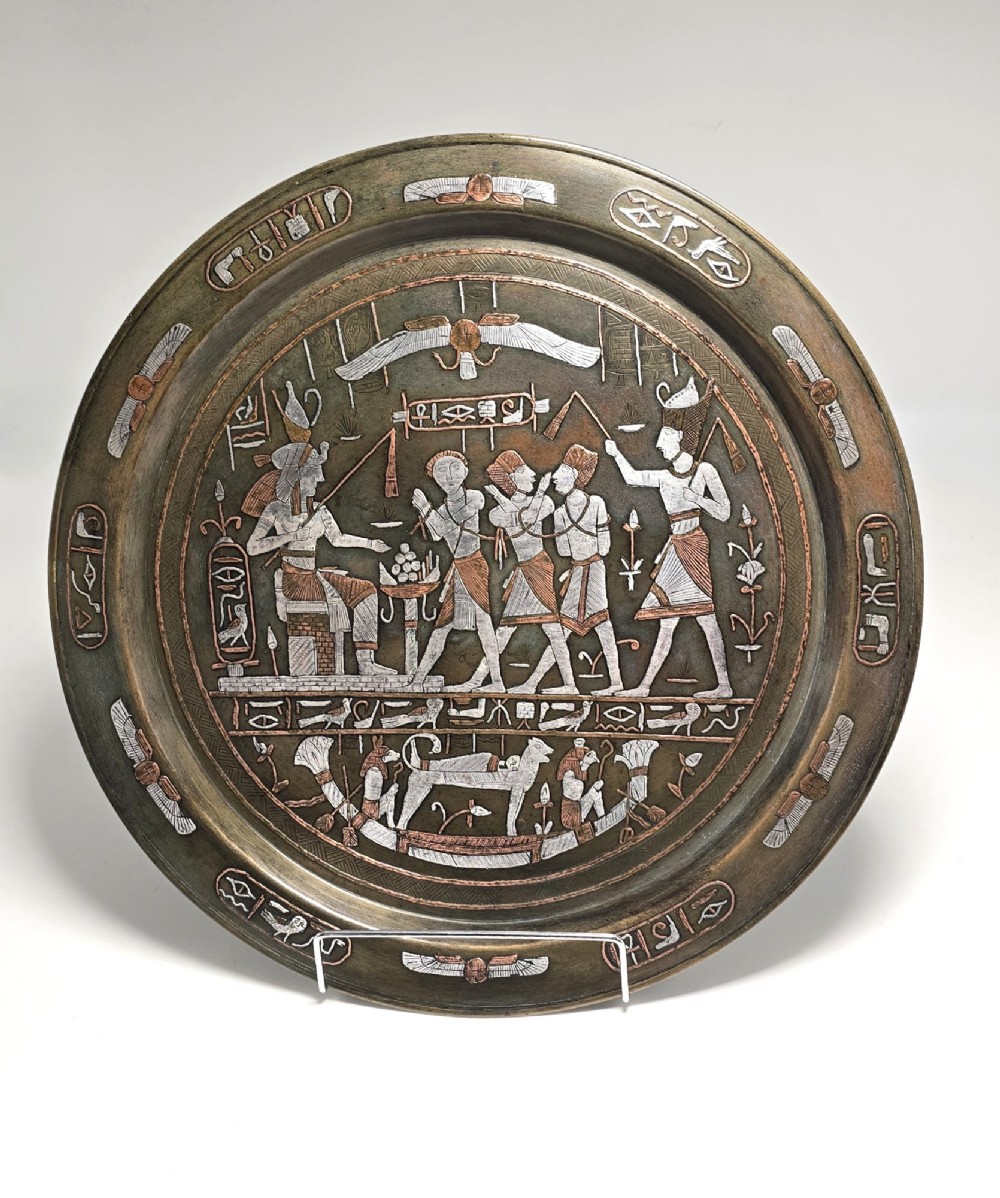 A circular bronze charger decorated with silver and copper inlays depicting pharaonic figures, hieroglyphs, and a winged sun deity.