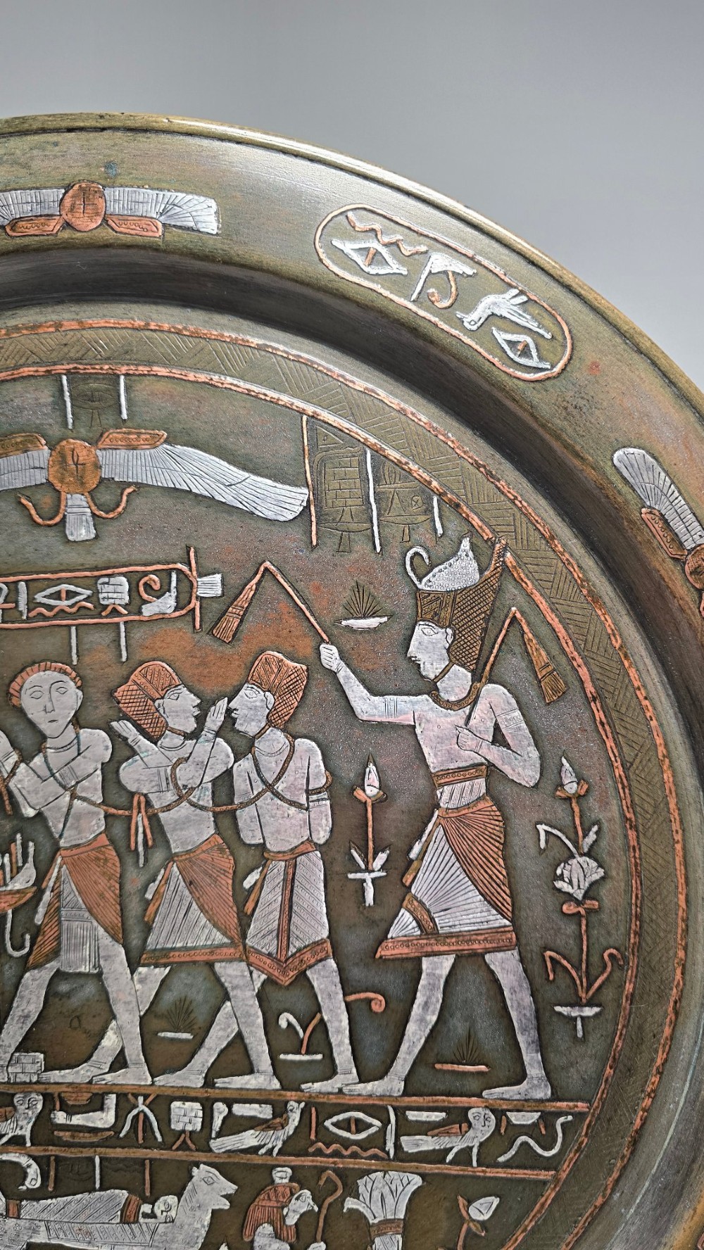 A circular bronze charger decorated with silver and copper inlays depicting pharaonic figures, hieroglyphs, and a winged sun deity. View III