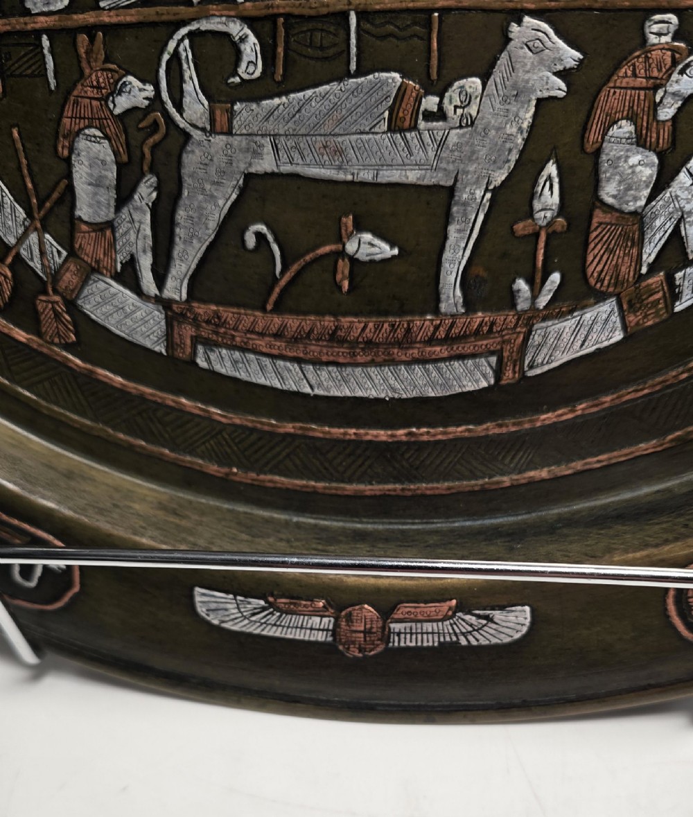 A circular bronze charger decorated with silver and copper inlays depicting pharaonic figures, hieroglyphs, and a winged sun deity. View VI
