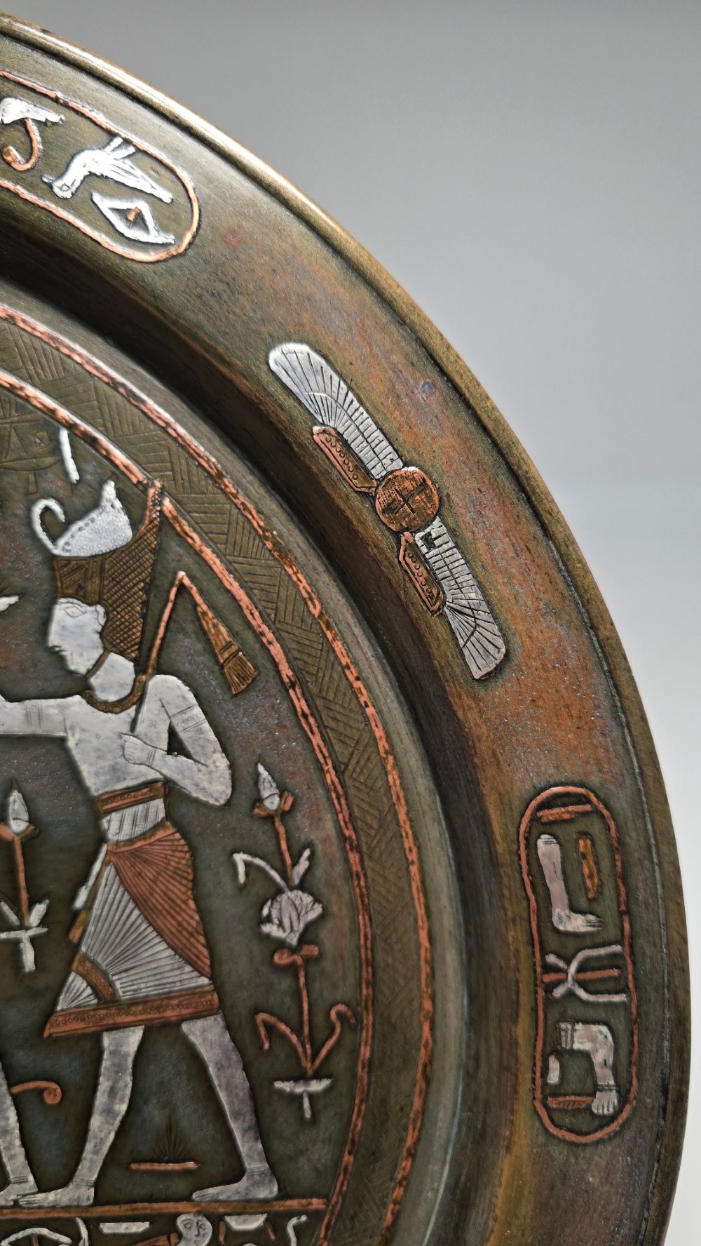 A circular bronze charger decorated with silver and copper inlays depicting pharaonic figures, hieroglyphs, and a winged sun deity. View VII