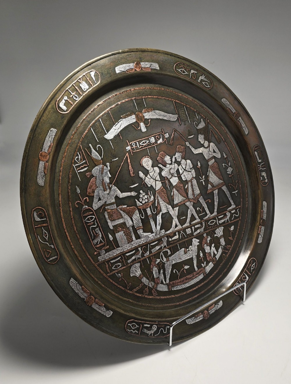A circular bronze charger decorated with silver and copper inlays depicting pharaonic figures, hieroglyphs, and a winged sun deity. View IX