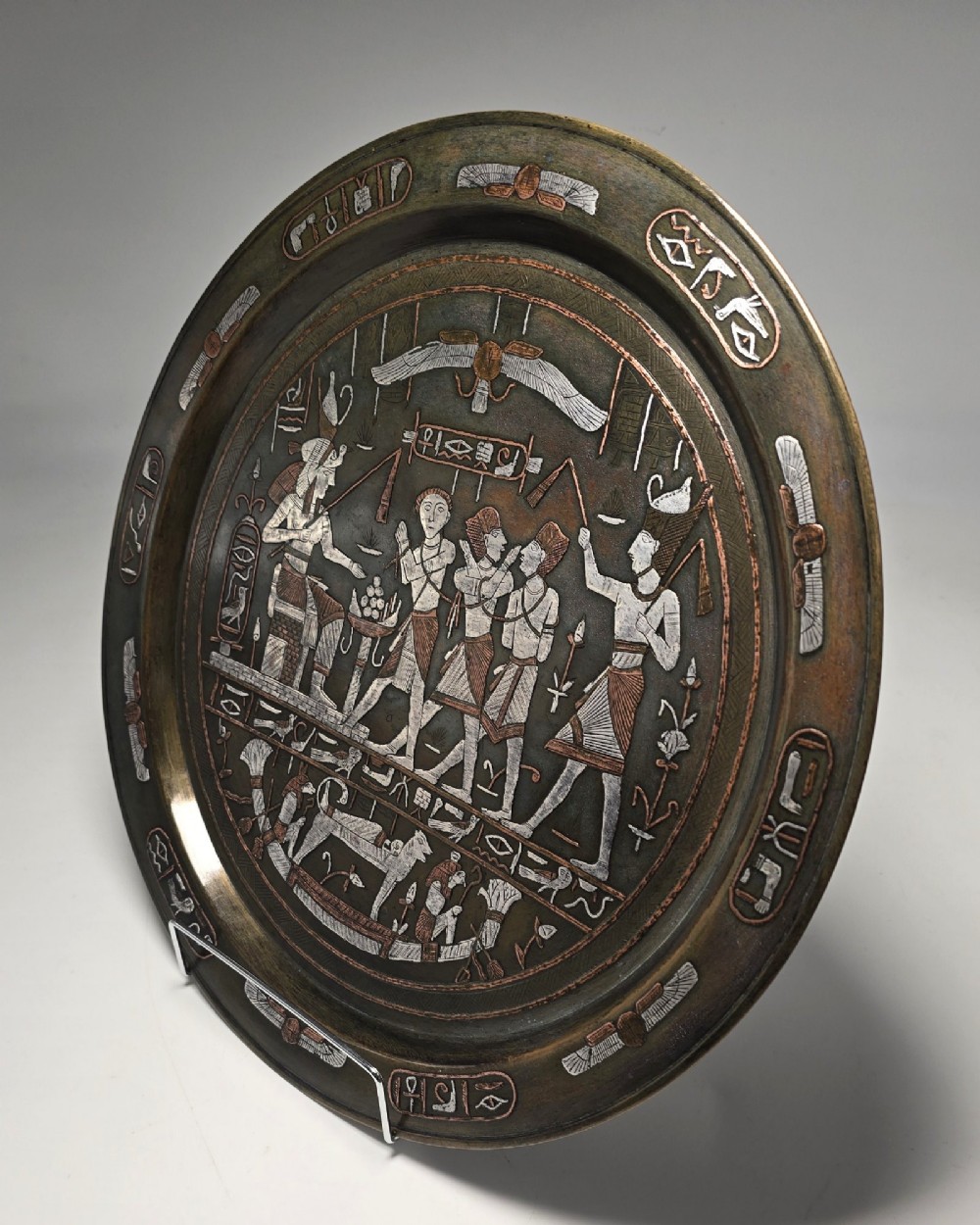 A circular bronze charger decorated with silver and copper inlays depicting pharaonic figures, hieroglyphs, and a winged sun deity. View X